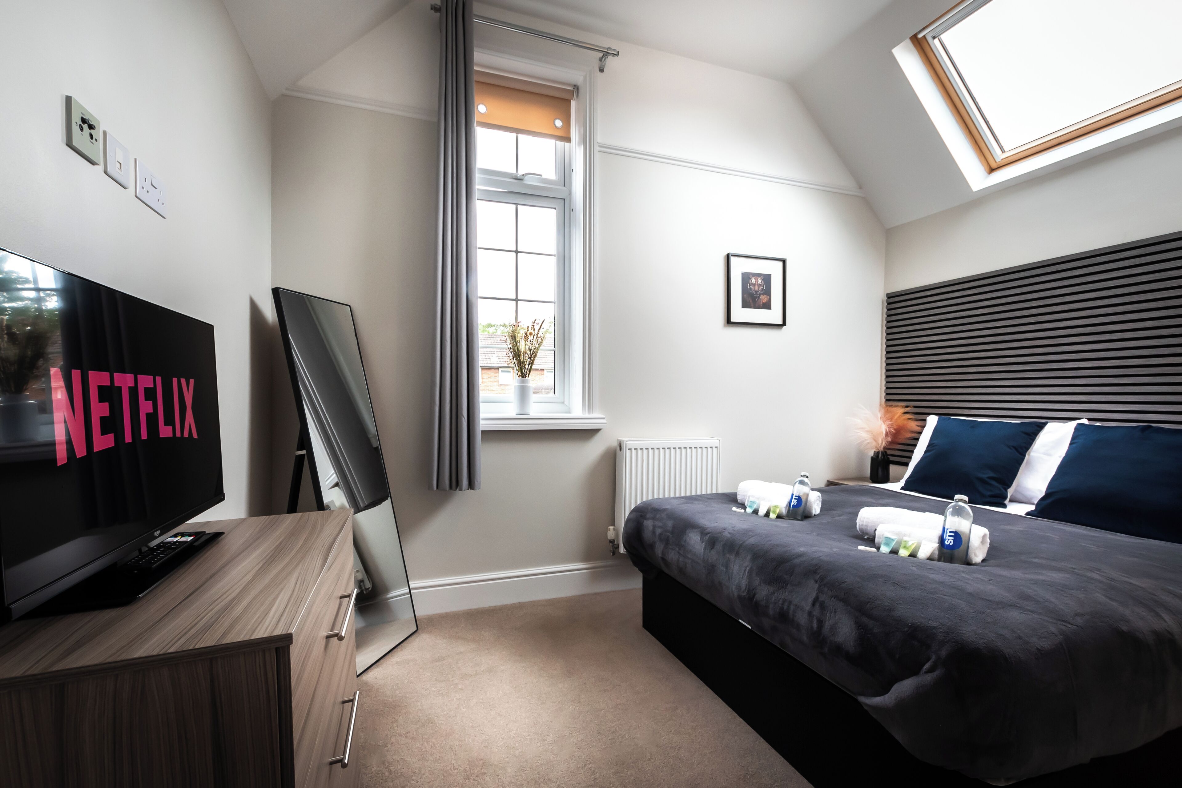 A modern hotel room with a flat-screen TV, a bed with a dark gray bedspread, a window with blinds, and a skylight.
