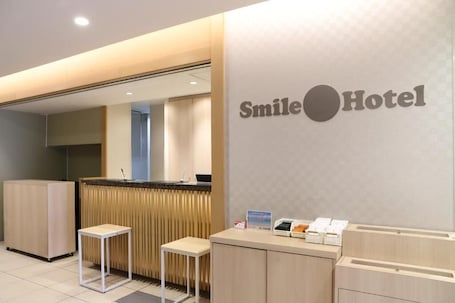 A reception area with a counter, two stools, a wall-mounted sign reading "Smile Hotel", and a small desk with a computer and brochures.. Smile Hotel Osaka Tennoji
