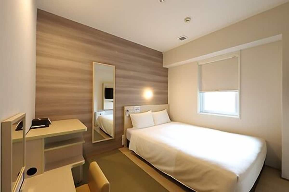 Une chambre d’hôtel avec un lit, un bureau, une chaise, un miroir et une fenêtre.