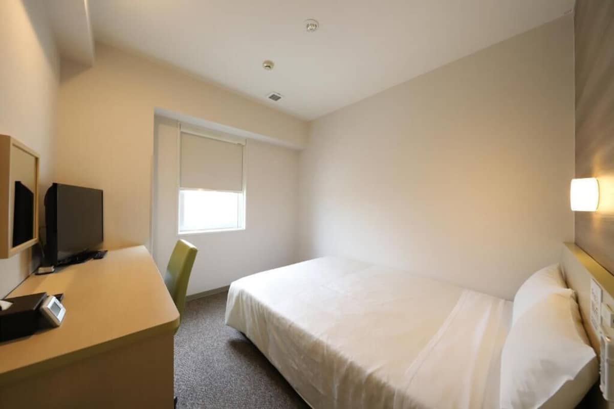 Une chambre d’hôtel avec un lit, un bureau équipé d’un ordinateur, une télévision et une fenêtre avec des stores.