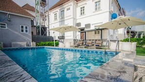 Una piscina con borde de piedra, un área de descanso con sillas y sombrillas, y un edificio de fachada blanca al fondo.