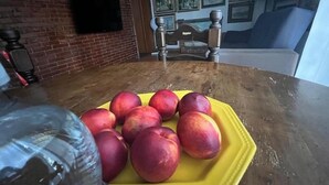 A dining table with a yellow plate of red peaches, a glass of water, and a brick wall with a TV mounted above it. - Eno 's house (Lucca)
