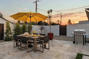 An outdoor patio with a table set for dining, a grill, and a large umbrella.