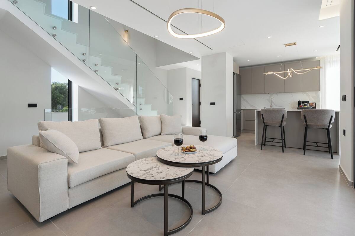 A modern living room with a beige sectional sofa, two round marble tables, and a staircase with glass railings.