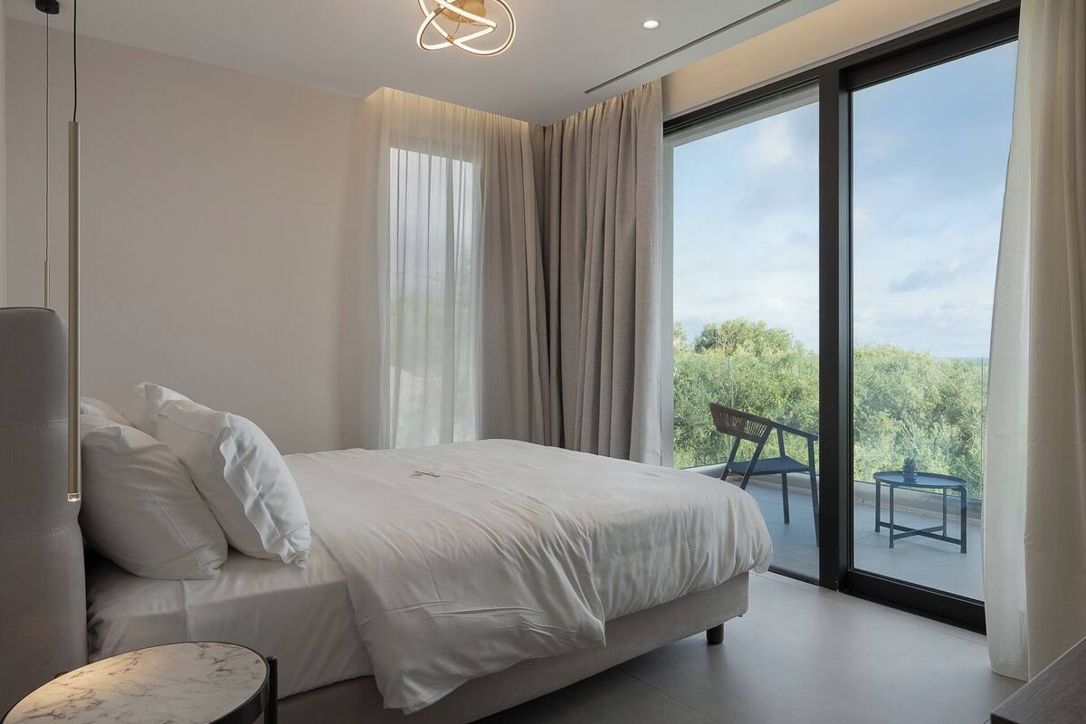 A modern bedroom with a large bed, a marble side table, and a view of greenery through a sliding glass door.