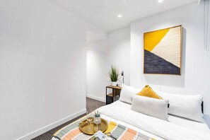Une chambre d’hôtel moderne avec un lit soigneusement fait, une petite table de chevet agrémentée d’une plante et un grand tableau abstrait accroché au mur.