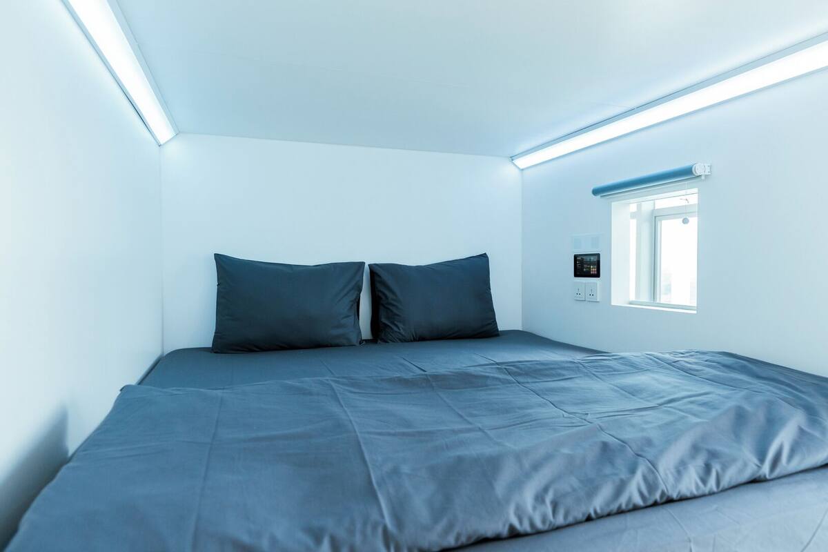 Een minimalistische slaapkamer met een enkel bed, een raam en een bedieningspaneel aan de muur.