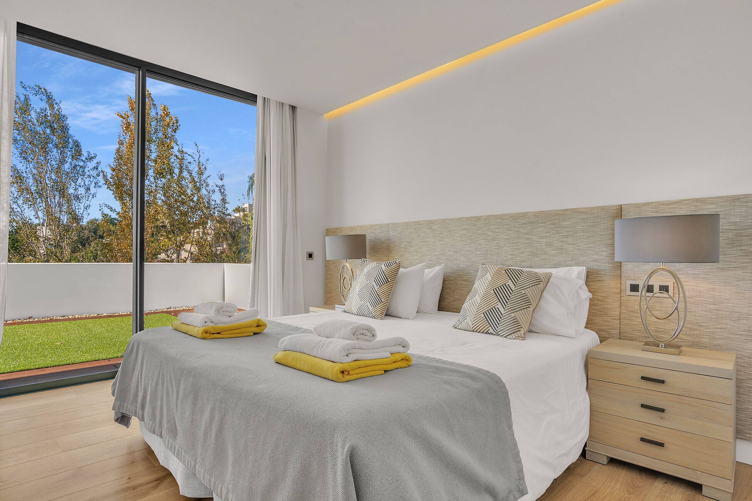 A modern bedroom with a large bed, a bedside table, and a view of a green lawn.
