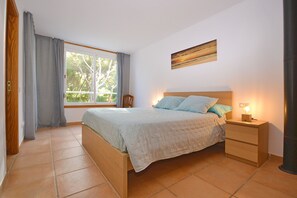 Ein Schlafzimmer mit einem Bett, einem Nachttisch, einem Sessel, einem Fenster mit Vorhängen und einem Gemälde an der Wand.