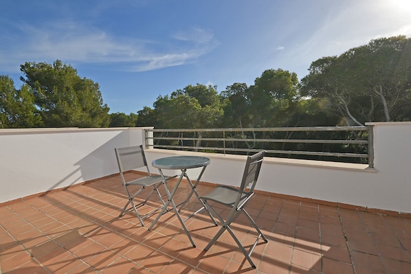 A terrace with a table and two chairs, a white wall, and a view of trees.