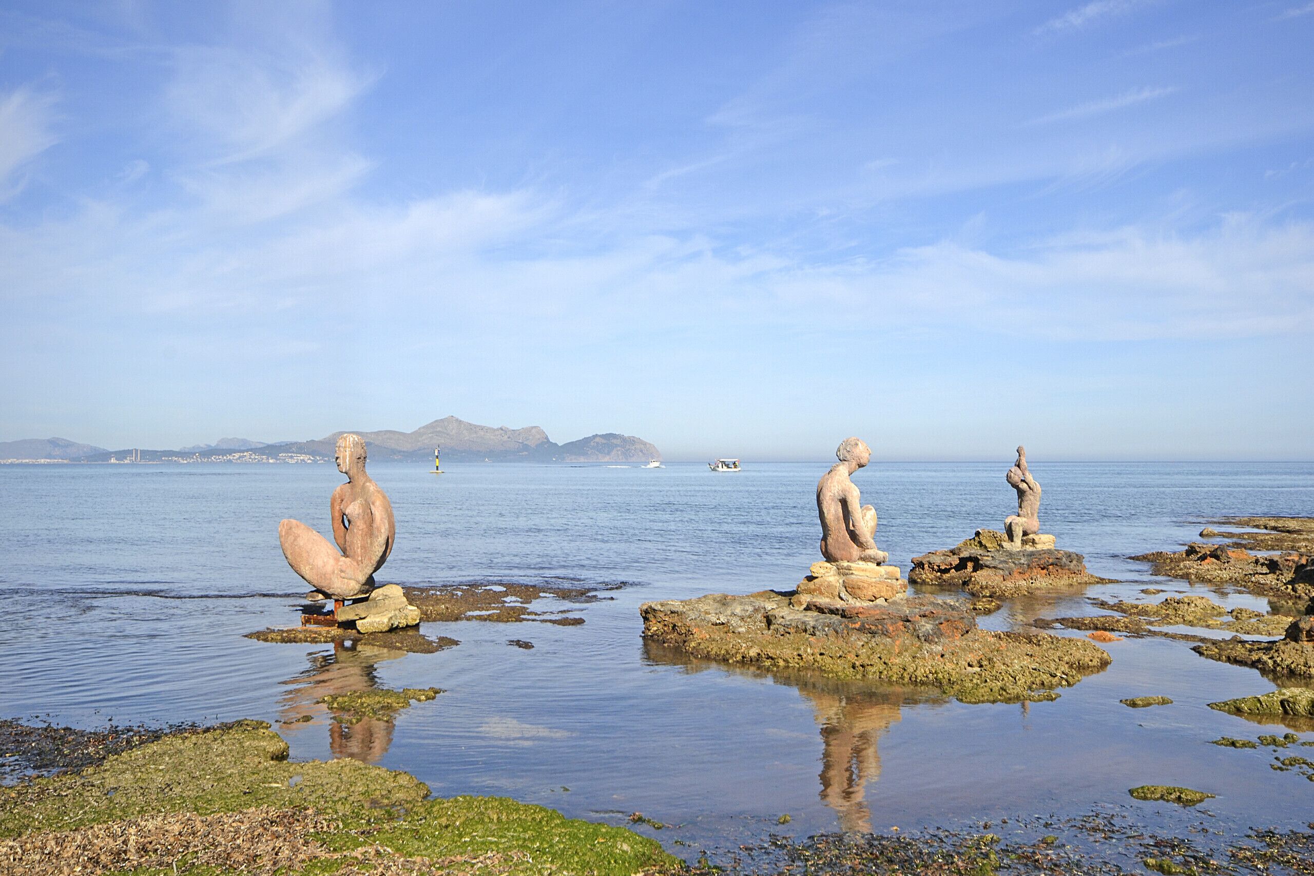 A coastal scene with three stone sculptures on rocks, calm water, and distant mountains.