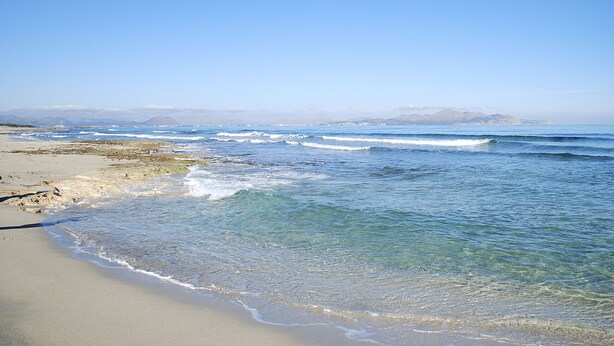 Ein Strand mit klarem Wasser, Wellen und Bergen in der Ferne.
