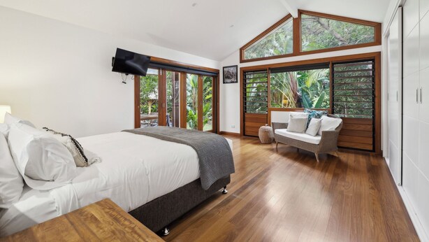 Een slaapkamer met een bed, een aan de muur bevestigde televisie, een bank en grote ramen met uitzicht op groen.