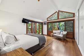 A bedroom with a bed, a TV mounted on the wall, a sofa, and large windows with a view of greenery. - Beached - Beach is your Back Yard, Pet Friendly (Sawtell)
