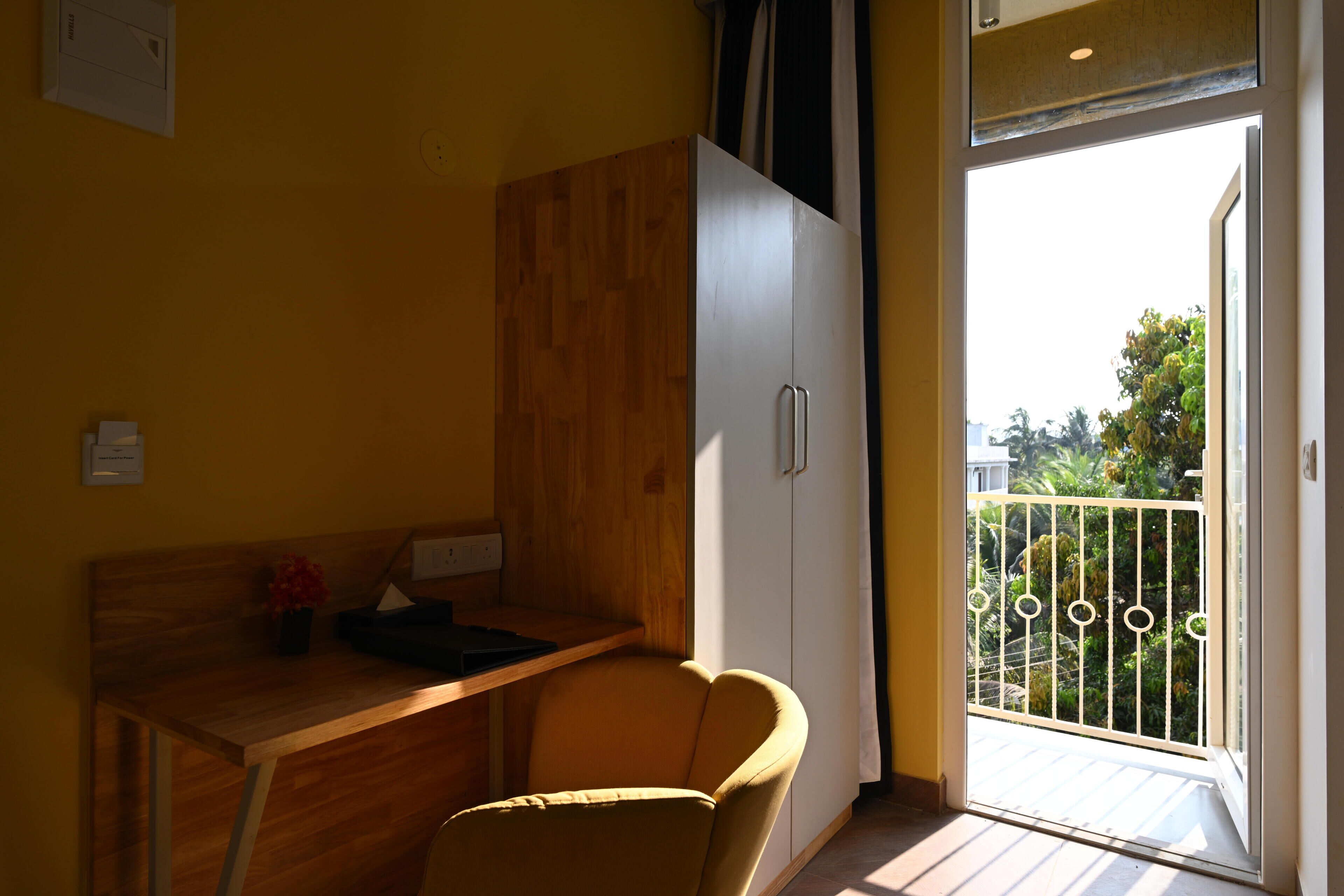 A room with a wooden desk, a chair, a white wardrobe, and a balcony with a metal railing.