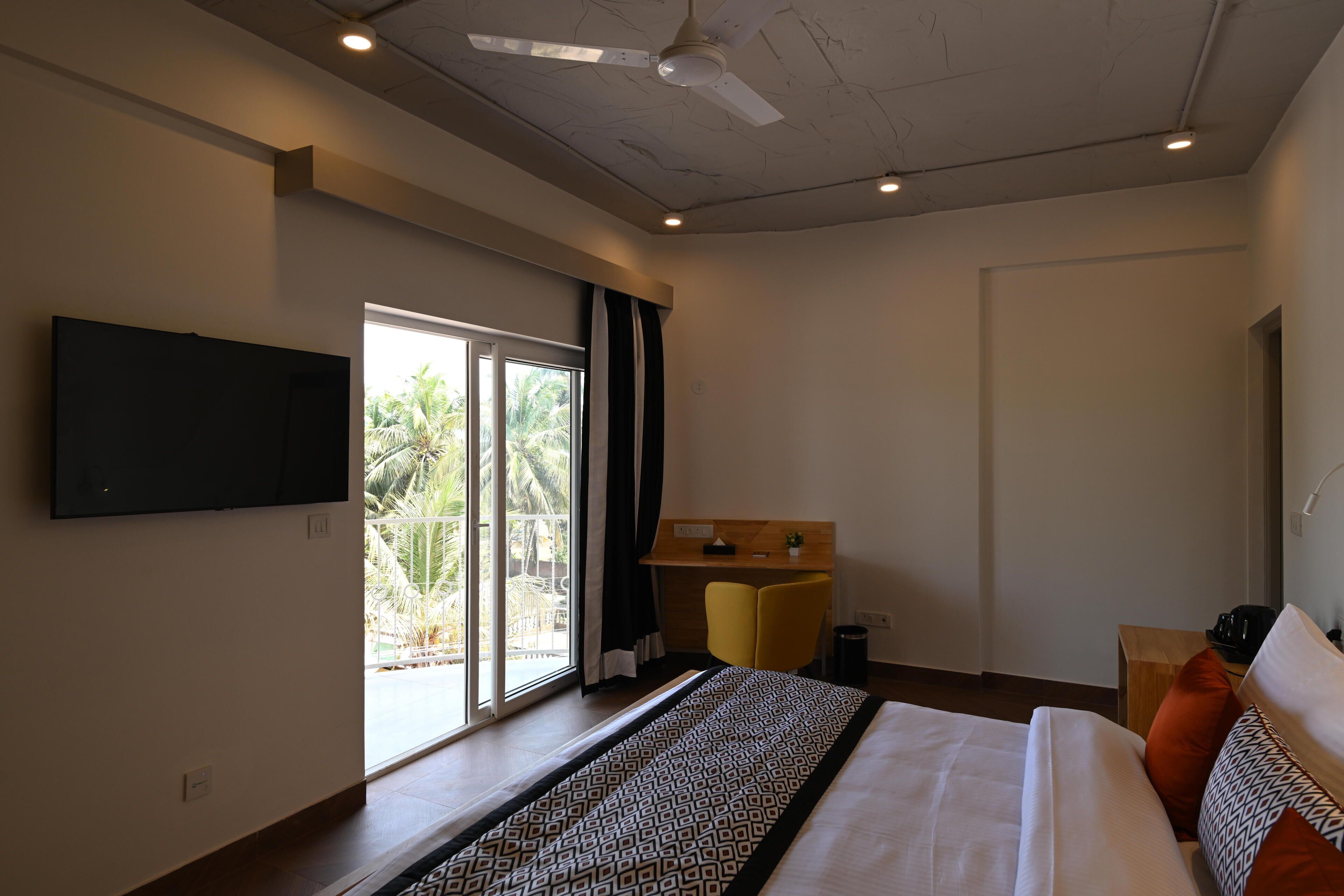 A modern hotel room with a large bed, a flat-screen TV mounted on the wall, a desk with a chair, and a balcony with a view of palm trees.