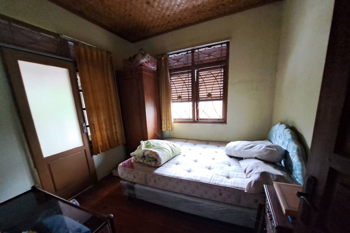 A small bedroom with a single bed, a wooden wardrobe, a window with blinds, and a door.