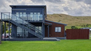 Ein modernes, zweistöckiges Haus mit einer Glasstufen-Treppe, einer Terrasse und Blick auf einen Hügel.