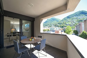 Un balcon avec une table et des chaises pour prendre les repas, une vue sur une ville située sur une colline, et un intérieur moderne agrémenté d’une lampe et d’une fenêtre.