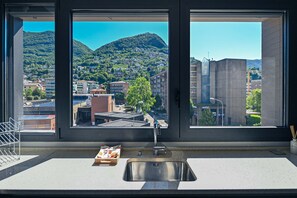 A kitchen with a large window, a stainless steel sink, and a view of a hillside town.