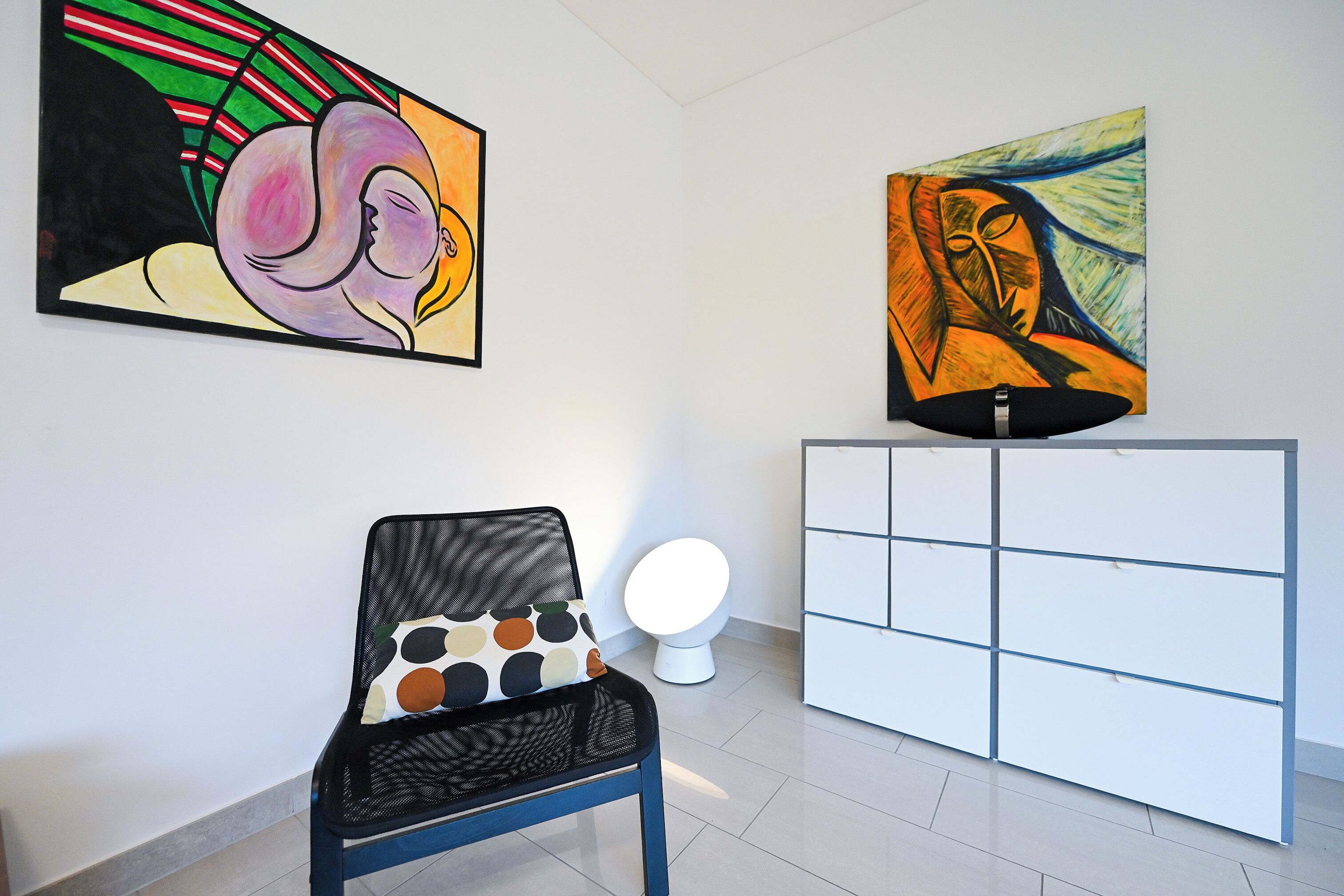A room with a chair, a white cabinet, and two paintings on the wall.