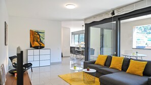A modern living room with a grey sofa, yellow cushions, a painting, and a dining area with a table and chairs. - Central Plaza Apartment 3 in Lugano (Lugano)