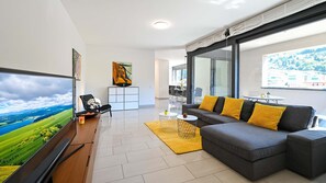 A modern living room with a large TV, a sofa with yellow cushions, a dining area, and a view of a green landscape. - Central Plaza Apartment 3 in Lugano (Lugano)