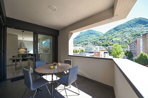 A balcony with a dining table and chairs, a view of a hillside town, and a modern interior with a lamp and a window.