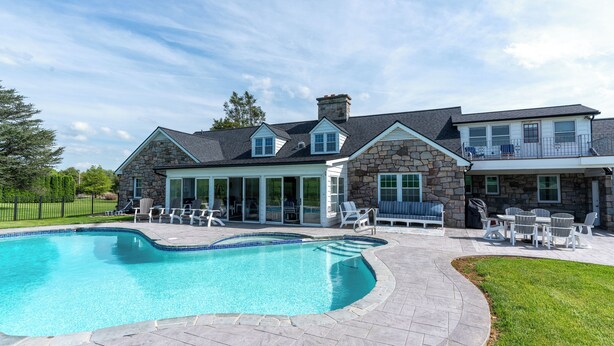Une grande piscine avec une terrasse en pierre, des espaces de détente en plein air et une maison en pierre dotée d’un balcon.