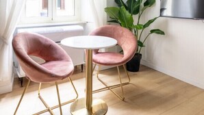 Une pièce avec un plancher en bois, une table ronde, deux chaises roses, une grande plante verte, un téléviseur accroché au mur et une fenêtre avec des rideaux blancs.