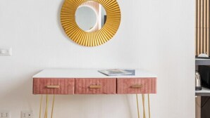 Un mur blanc orné d’un miroir à bordure dorée, une console rose avec des tiroirs et des pieds dorés.