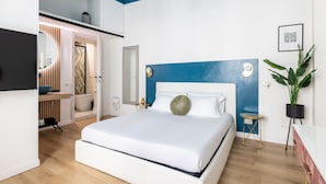 Une chambre moderne avec un grand lit, un mur d’accent bleu, une petite table de chevet, une plante en pot et un téléviseur fixé au mur.