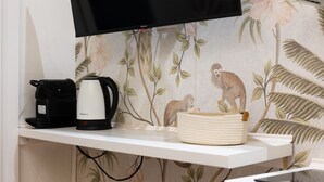 A kitchen with a flat-screen TV mounted above a white countertop, a coffee maker, a stack of plates, and two pink bar stools. - Cozy Love Nest Steps from Colosseum (Roma)