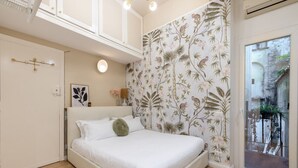 A bedroom with a bed, a wall with a floral pattern, a door with a light switch, and a window with a view of the outside. - Cozy Love Nest Steps from Colosseum (Roma)