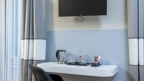 A modern hotel room with a flat-screen TV mounted on the wall, a small desk with a kettle, glasses, and a snack tray, a dark chair, and wooden flooring. - Tiberia Room - Suites Romae Rex (Roma)