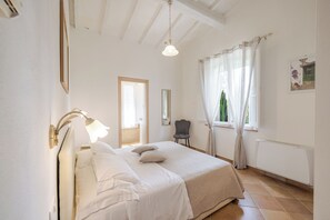 A bedroom with a bed, a chair, a window with curtains, a radiator, and a light fixture.