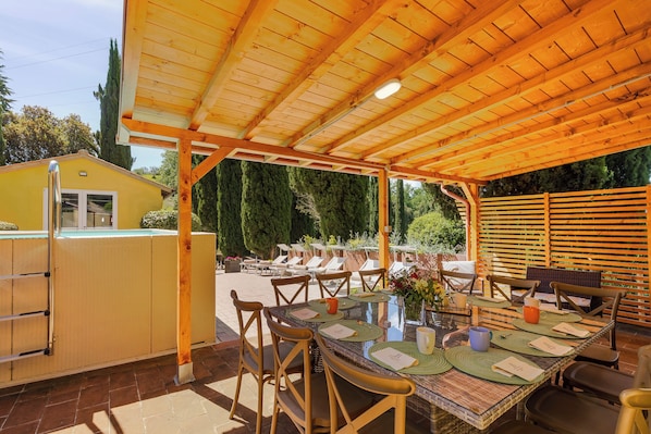 An outdoor patio with a wooden pergola, a dining table set with chairs, and a yellow building in the background.