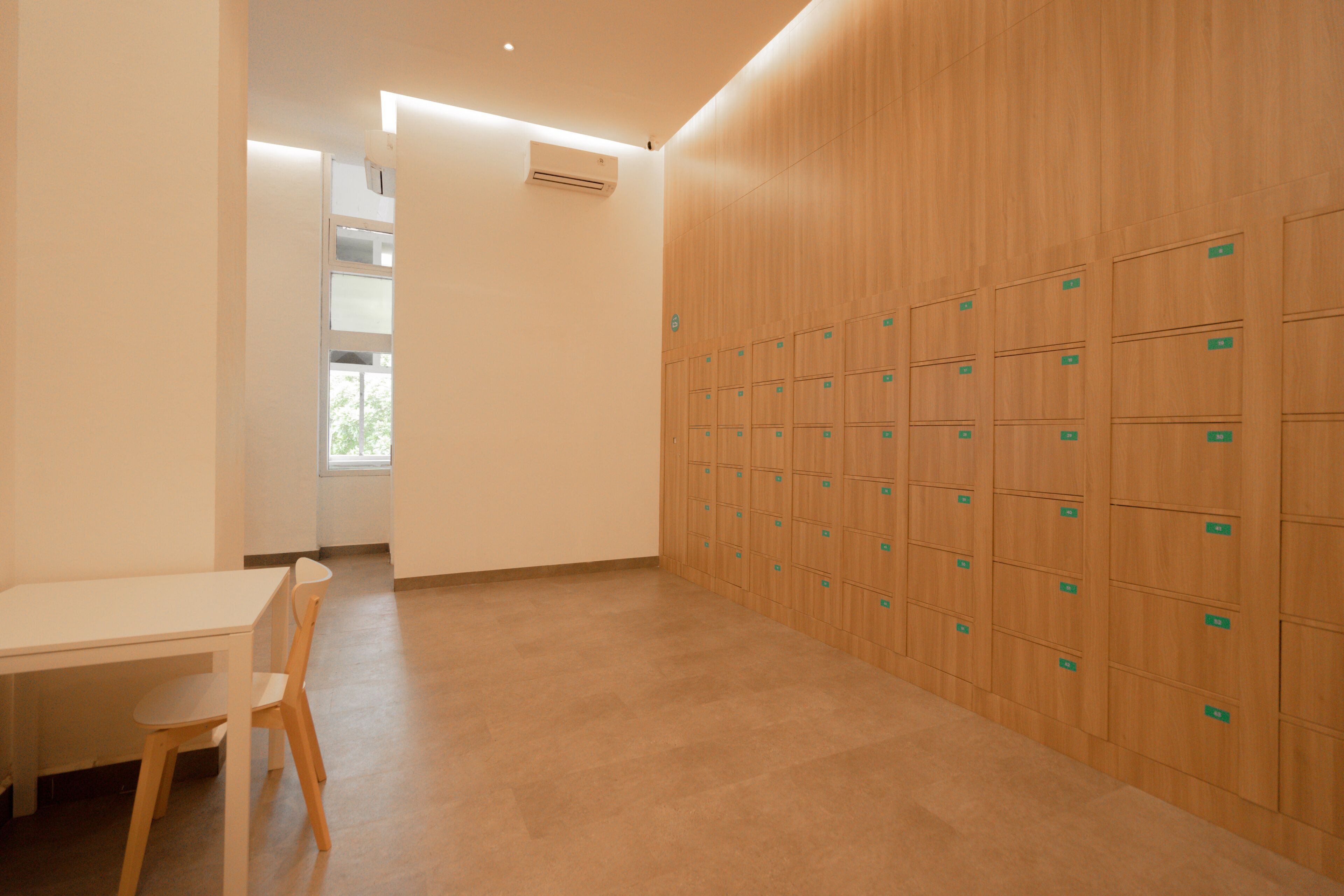 Une pièce avec un mur composé de casiers en bois, une table blanche avec une chaise et une fenêtre donnant sur des arbres.