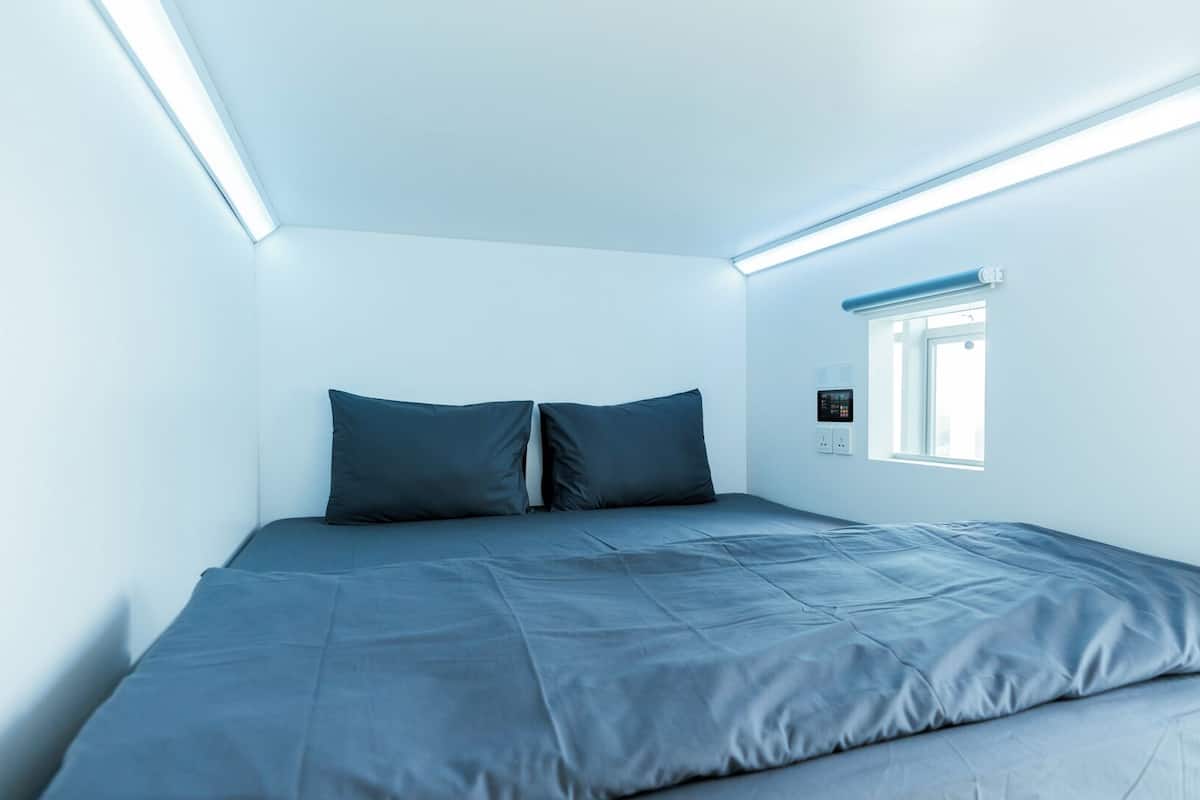 A small, modern bedroom with a single bed, two pillows, and a window with blinds.