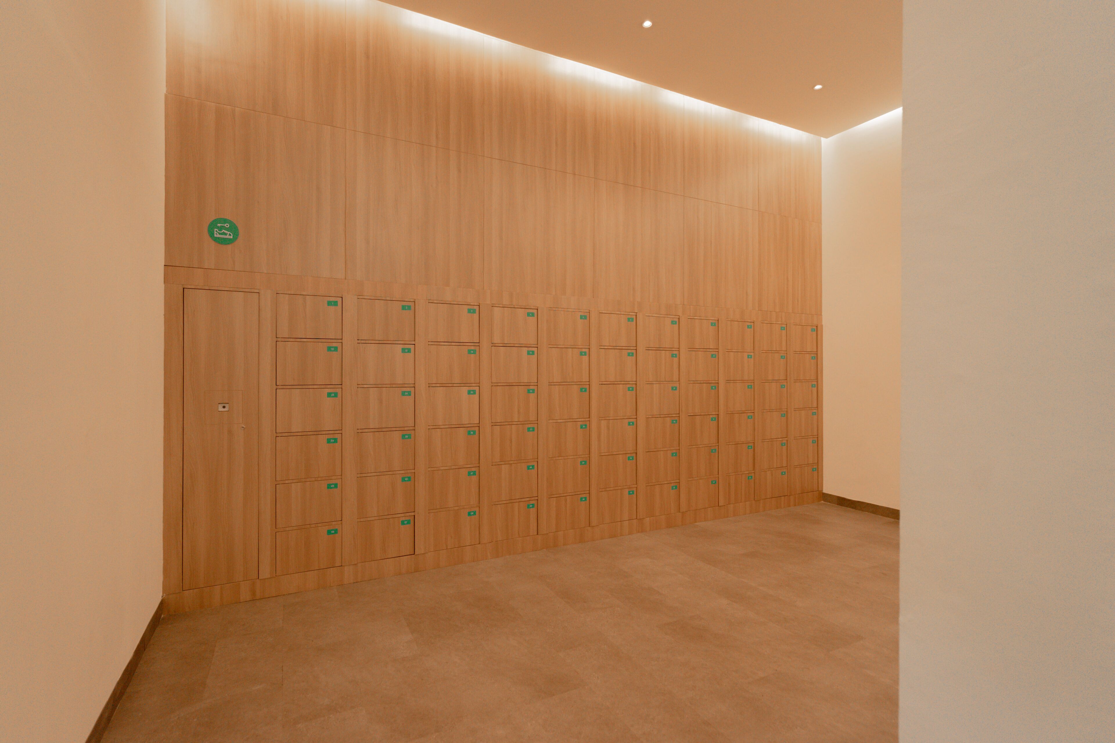 A row of wooden lockers with green labels, a beige carpeted floor, and a plain white wall.