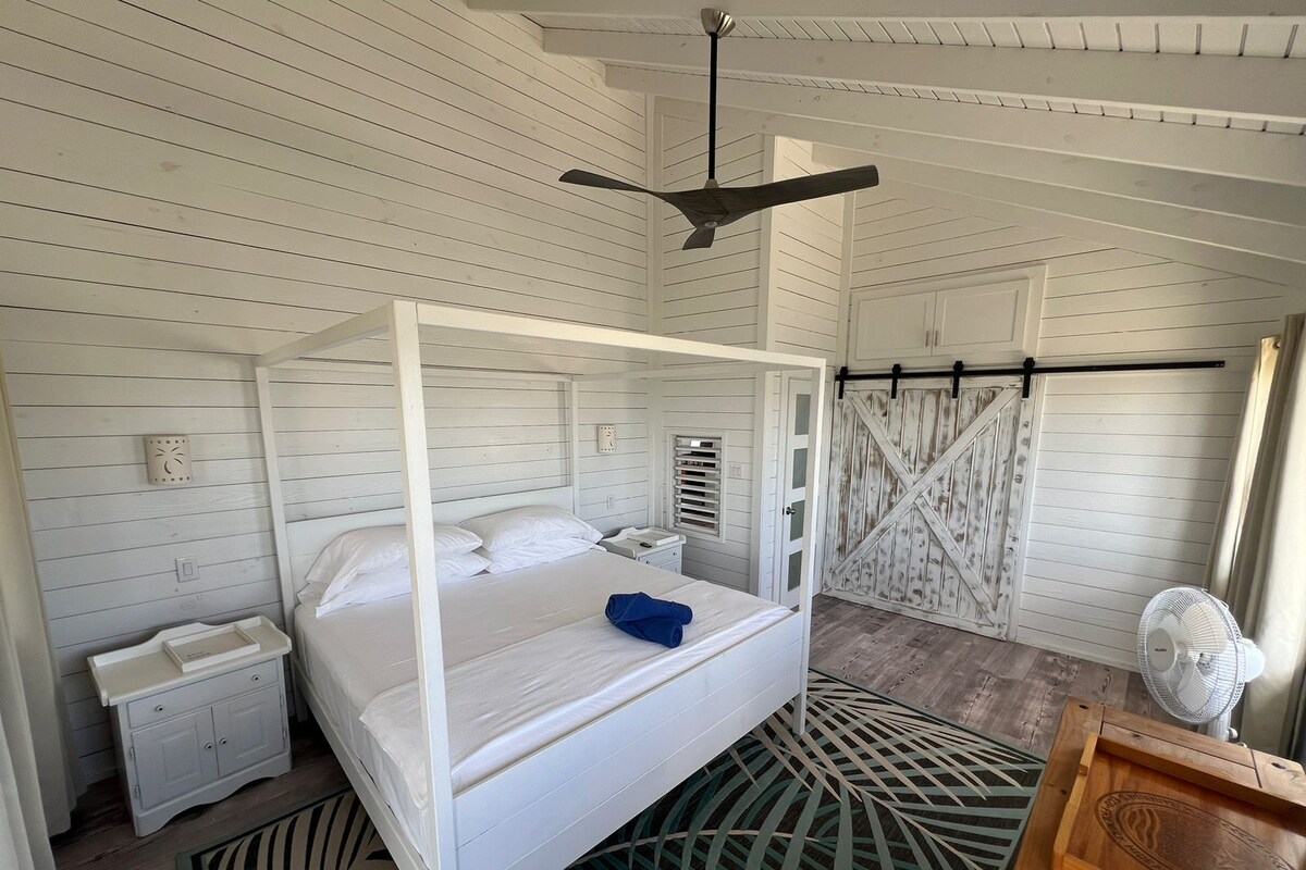 A white four-poster bed with a blue towel, a wooden nightstand, a white wall clock, and a rustic barn-style door.