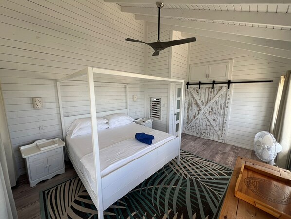 A white four-poster bed with a blue towel, a wooden nightstand, a white wall clock, and a rustic barn-style door. - Barbuda Cottages (Codrington)