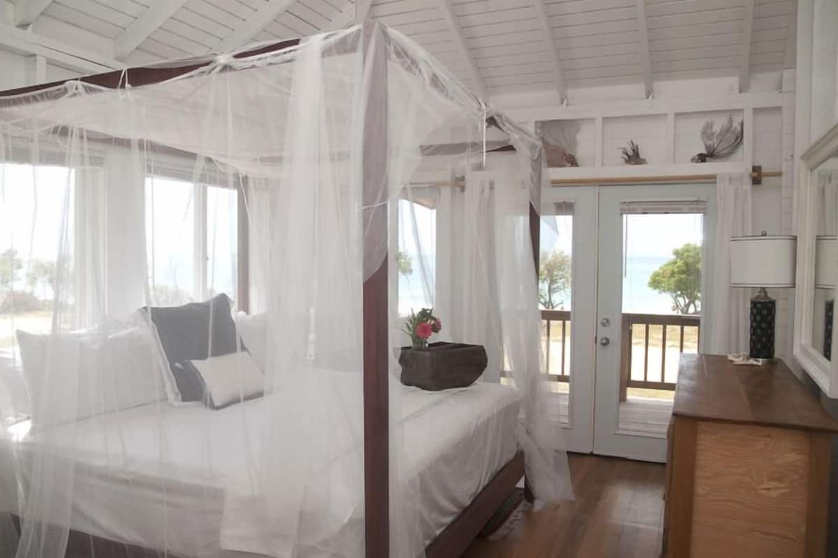 A bedroom with a four-poster bed, a wooden dresser, and a view of the ocean through the door.
