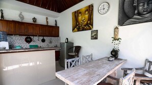 Une cuisine avec des meubles de rangement en bois, une table à manger, des chaises, un réfrigérateur et des éléments de décoration murale, notamment un tableau représentant Bouddha et une horloge.