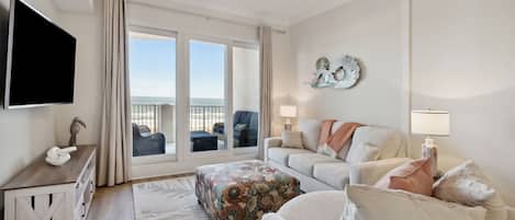 A spacious living room with a large window, a ceiling fan, a flat-screen TV, a sofa, a chair, and a decorative wall piece.
