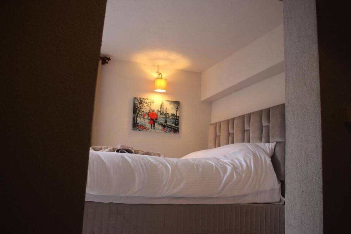 A single bed with a headboard, a wall-mounted light, and a painting of a cityscape.