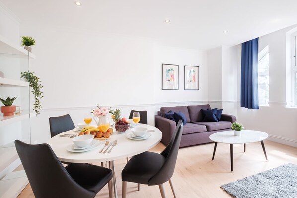 A modern living room with a dining area. There is a round dining table with chairs, a sofa, and a coffee table. The room has a large window with curtains, a plant on the windowsill, and artwork on the wall.