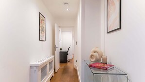 A hallway with a radiator, a glass table with books and a candle, and framed artwork on the walls. - 2BR 2BA near Hyde Park & Kensington (London)