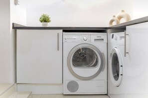 A compact laundry room with a front-loading washing machine and dryer, a small potted plant, and a white countertop. - 2BR 2BA near Hyde Park & Kensington (London)