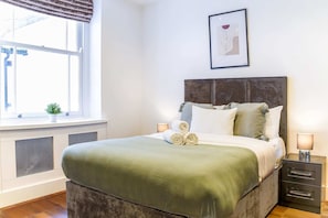 Une chambre à coucher bien rangée, avec un lit, des lampes de chevet, une fenêtre donnant sur un beau paysage et une œuvre d’art encadrée au mur.